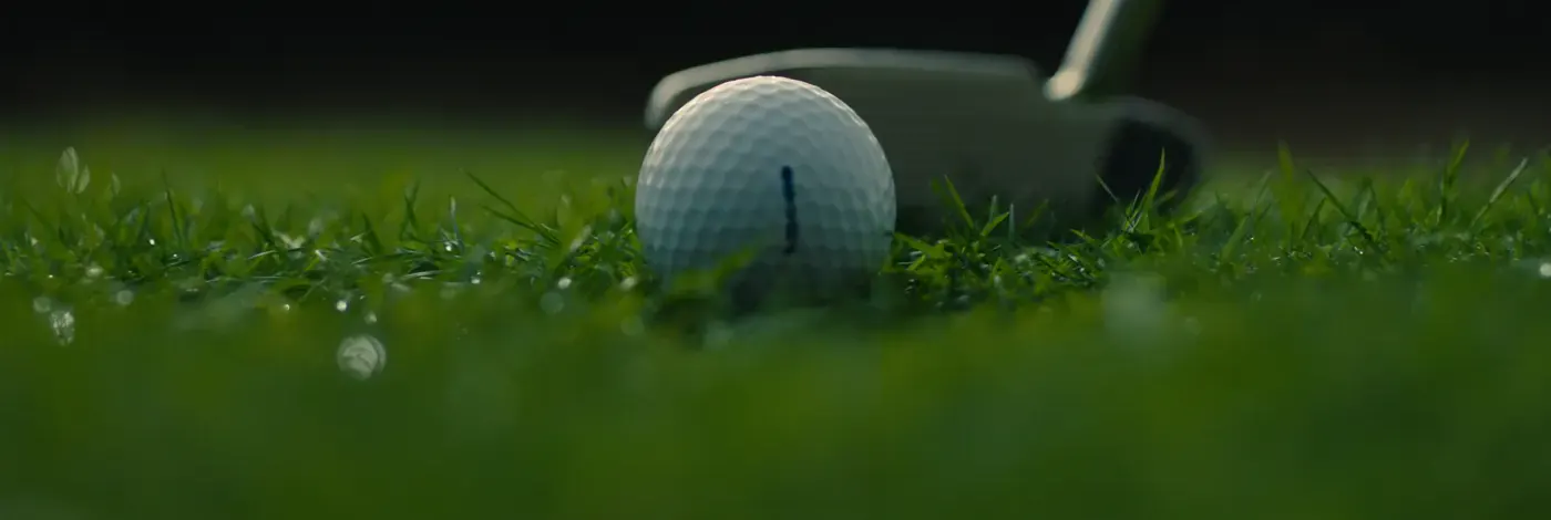 A golf ball on dewy short grass beside the head of a putter